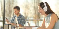 A lady with headphones looking and smiling at the screen of her laptop. There's a guy on the background looking at his smartphone.