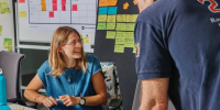 A teacher on her desk, smiling while talking to a colleague. There's a blackboard with sticky notes behind them.
